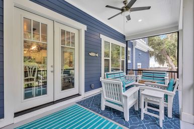 Coastal-style screened porch with navy blue siding, white French doors and ceiling fan, featuring white patio chairs and sofa with teal-striped cushions on patterned rugs.