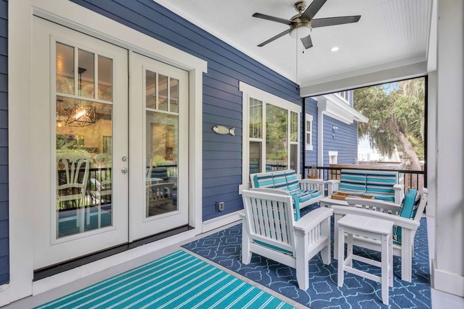 Coastal-style screened porch with navy blue siding, white French doors and ceiling fan, featuring white patio chairs and sofa with teal-striped cushions on patterned rugs.