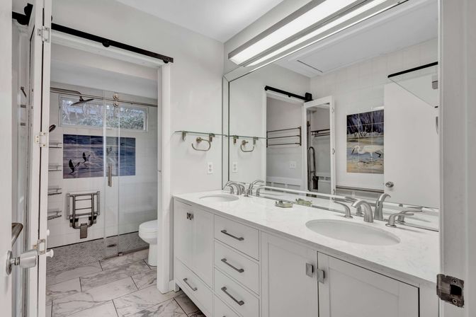 Bright modern master bathroom with white double-sink marble vanity, large mirror, glass walk-in shower with blue bird ocean tile mural and marble floor tiles