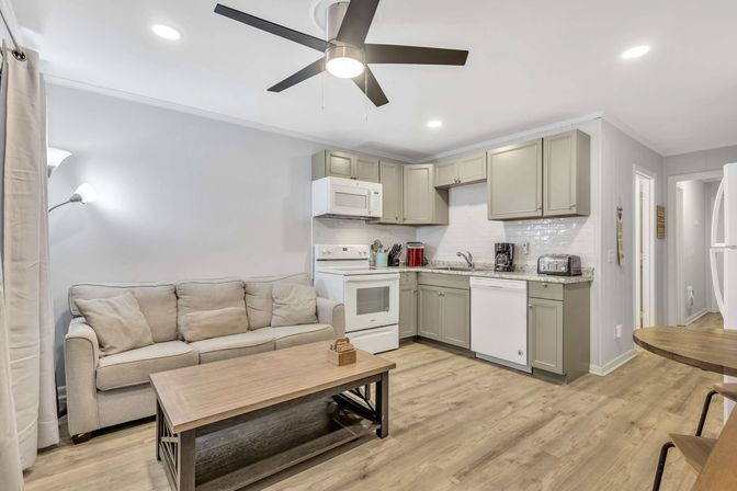 Cozy open-plan living room and kitchen in a bright rental: beige sofa, wooden coffee table, gray cabinets, white appliances, ceiling fan, wood-look floors