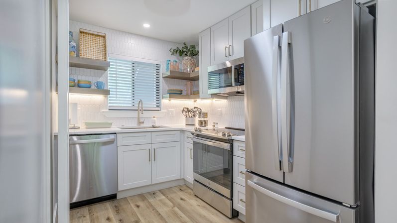 Bright modern L-shaped kitchen with white shaker cabinets, stainless-steel French-door refrigerator and appliances, open wood shelves, hex-tile backsplash, wood-look flooring and window above the sink.