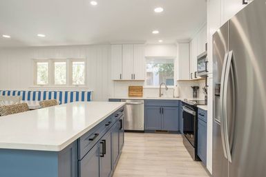 Bright coastal-inspired modern kitchen with large white quartz island, blue lower cabinets, white upper cabinets, stainless steel appliances, and a blue-and-white striped banquette