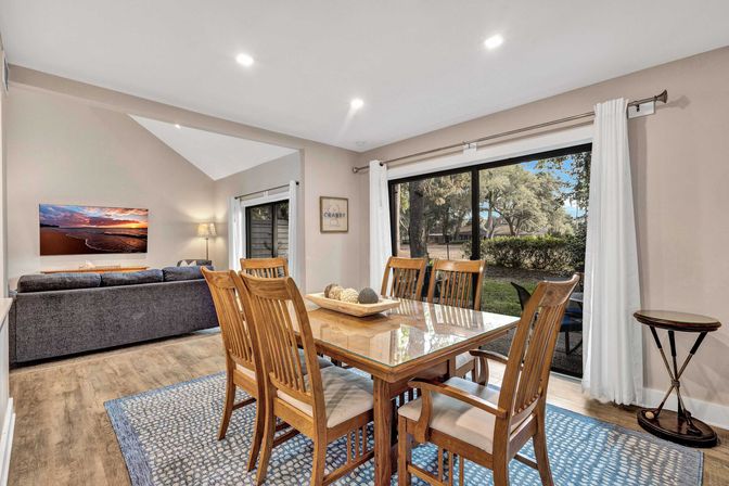Open-plan dining and living area with a wooden six-seat dining table on a blue rug, gray sofa, wall-mounted TV, hardwood floors, recessed lighting, and large sliding glass doors opening to a tree-lined backyard.