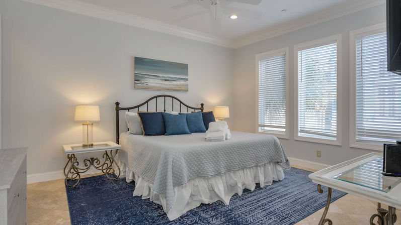 Cozy sunlit coastal-style bedroom with king bed and iron headboard, gray quilt, navy throw pillows, matching bedside lamps, coastal artwork above the bed, three tall windows with white blinds, and a navy area rug.