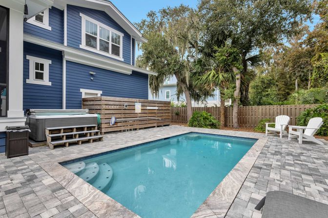 Inviting backyard rectangular swimming pool with built-in steps, gray paver patio, adjacent hot tub, two white Adirondack chairs, blue two-story house, palm trees and wooden fence.