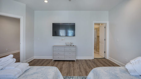 Bright airy bedroom with two beds and folded towels, wall-mounted flat-screen TV above a light wood dresser, hardwood floors, light gray walls and an open ensuite bathroom