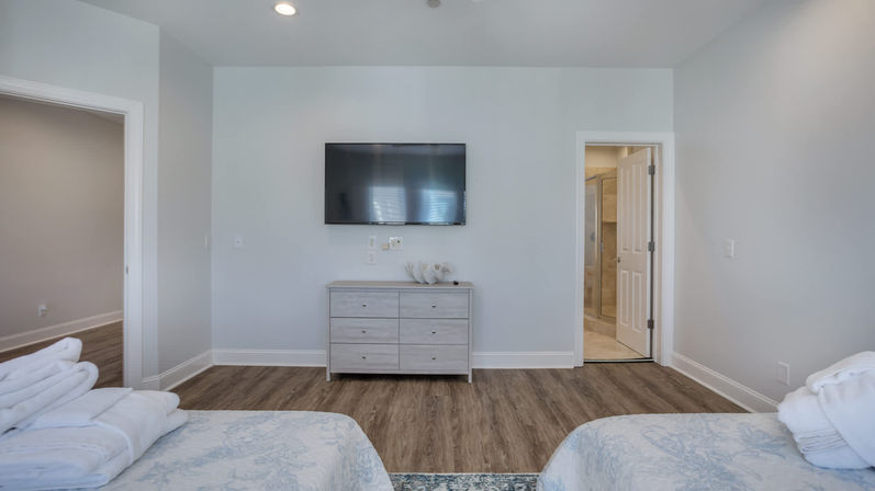 Bright airy bedroom with two beds and folded towels, wall-mounted flat-screen TV above a light wood dresser, hardwood floors, light gray walls and an open ensuite bathroom