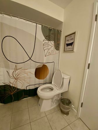 Bright residential bathroom with a white toilet (lid up), abstract botanical shower curtain, beige tiled floor, small trash can and framed humorous wall art.