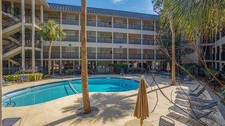 Sunlit kidney-shaped courtyard pool at a three-story hotel-style building with palm trees, blue lounge chairs, and a closed beige umbrella.