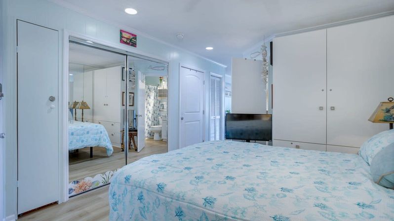 Bright beachy bedroom with sea‑foam patterned bedspread, mirrored sliding closet doors reflecting a bathroom, white built-in cabinets, small flat-screen TV, and light wood floors