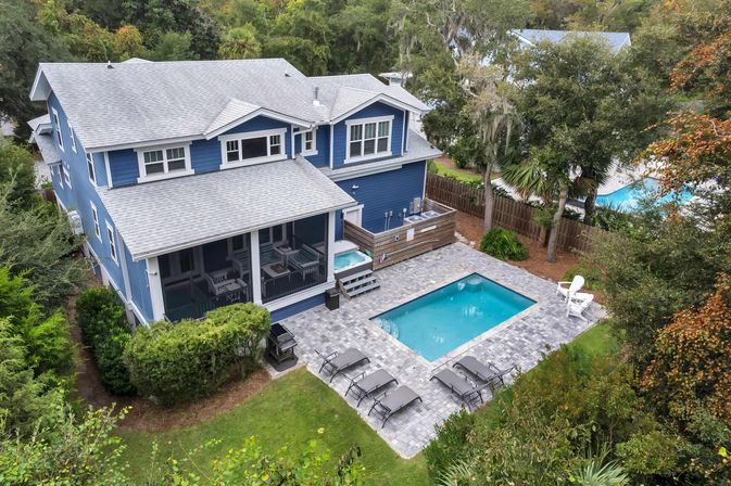Aerial view of a blue two-story coastal-style home with a screened porch, paver patio, built-in spa and rectangular backyard pool surrounded by lounge chairs, fencing and lush trees.