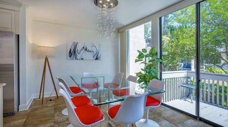 Bright modern dining room with a glass-top table, white chairs with red cushions, bubble pendant light and tripod floor lamp, opening through sliding glass doors to a sunny balcony with green trees.
