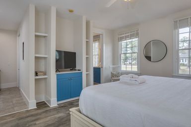 Bright, coastal-style bedroom with white walls, large windows, round mirror, queen bed with folded towels, blue built-in cabinet and TV, open shelving, and wood‑look floors.