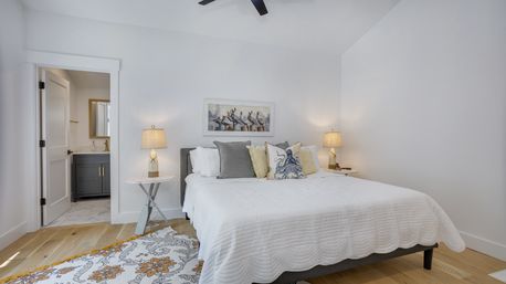 Bright modern coastal bedroom with white walls, hardwood floors, beachy pillows and pelican art, bedside lamps, ceiling fan and visible en-suite bathroom.
