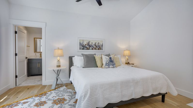 Bright modern coastal bedroom with white walls, hardwood floors, beachy pillows and pelican art, bedside lamps, ceiling fan and visible en-suite bathroom.