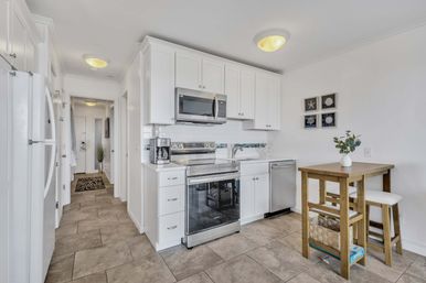 Bright, compact coastal-style kitchen with white cabinets, stainless-steel range and microwave, subway-tile backsplash, tiled floor, and a small wooden breakfast table with stools.