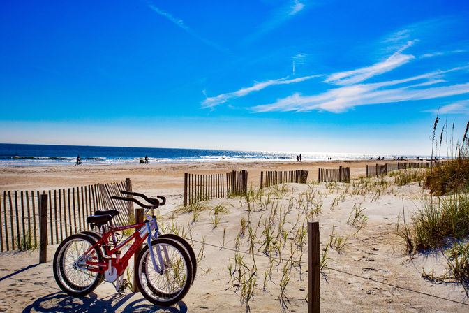 Sunny oceanfront beach with a red bicycle leaning against a wooden dune fence, sandy dunes with sea oats, sparkling blue water and people walking along the shoreline under a clear sky.