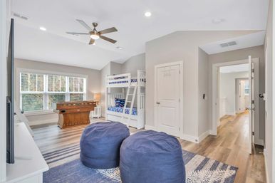 Bright, play-ready kids' game room with white bunk beds, wooden foosball table by a large window, two navy bean bags on a blue rug and wood-look floors.