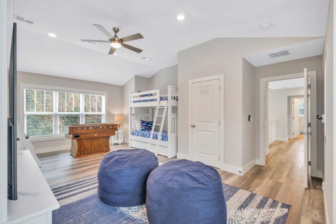 Bright, play-ready kids' game room with white bunk beds, wooden foosball table by a large window, two navy bean bags on a blue rug and wood-look floors.