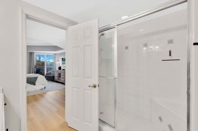 Bright master suite with en-suite white-tiled glass shower, open door to a bedroom with hardwood floors, bed, dresser and large window overlooking trees and a balcony.