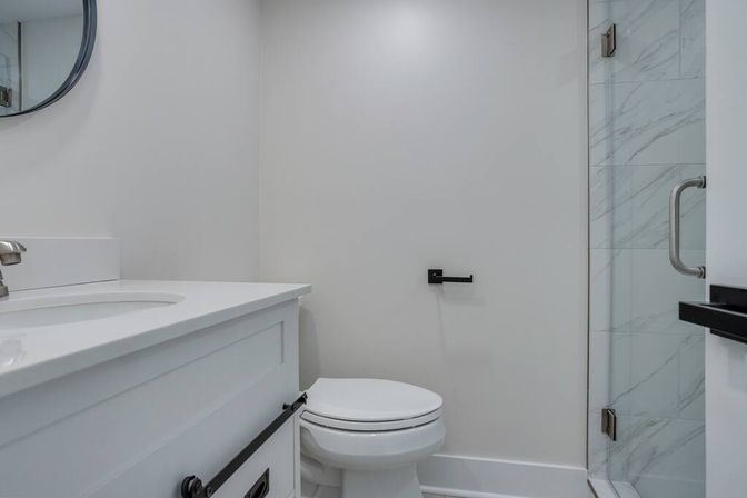 Bright compact modern bathroom with white vanity and sink, round mirror, white toilet, glass shower with marble-look tile, and matte black hardware.