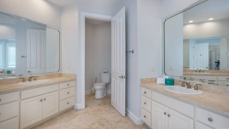 Bright modern master bathroom with double-sink vanity, marble countertops, large beveled mirrors, tile floor, chrome faucets, and an open door revealing a water closet.