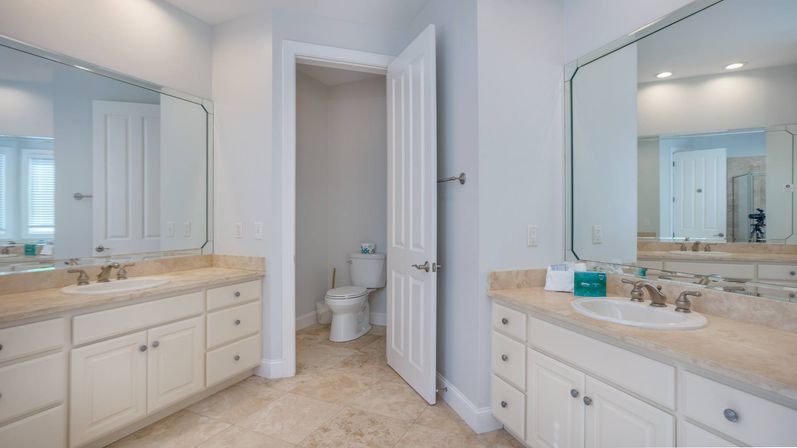 Bright modern master bathroom with double-sink vanity, marble countertops, large beveled mirrors, tile floor, chrome faucets, and an open door revealing a water closet.