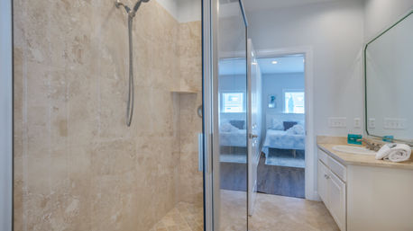 Bright modern ensuite bathroom with glass-framed walk-in shower in beige stone tile, single-sink vanity with mirror, and doorway revealing a light-filled bedroom