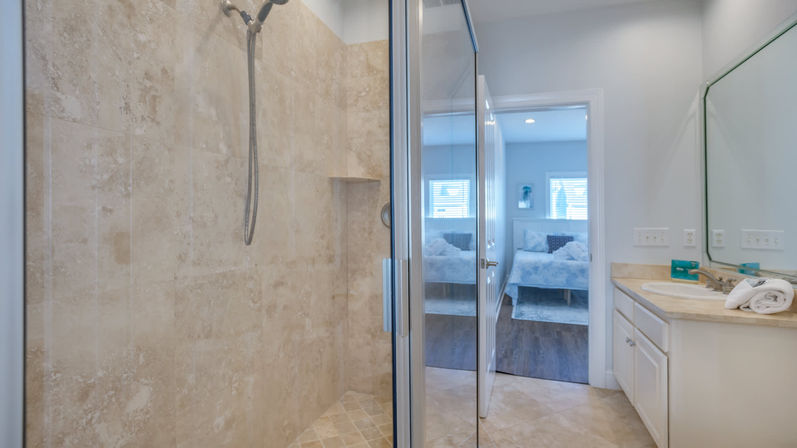 Bright modern ensuite bathroom with glass-framed walk-in shower in beige stone tile, single-sink vanity with mirror, and doorway revealing a light-filled bedroom