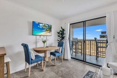 Bright beachfront condo dining nook with round wooden table, two blue upholstered chairs, wall-mounted TV, potted plant, and sliding glass doors opening to a balcony with bar stools and ocean view