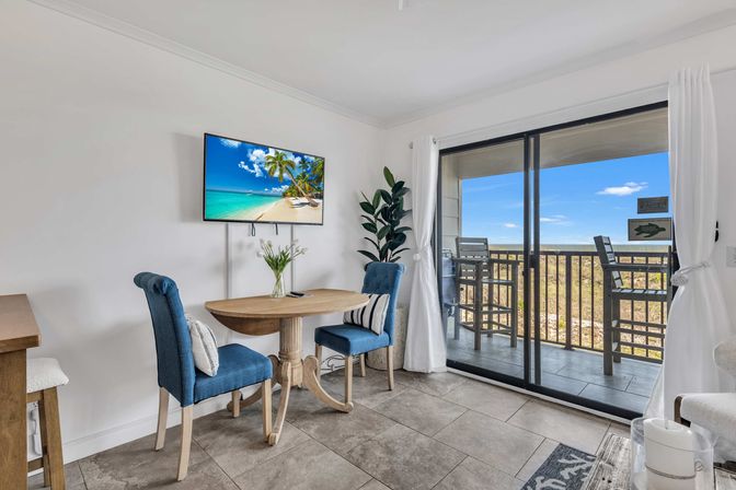 Bright beachfront condo dining nook with round wooden table, two blue upholstered chairs, wall-mounted TV, potted plant, and sliding glass doors opening to a balcony with bar stools and ocean view