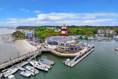Aerial view of a coastal marina with a red-and-white lighthouse, waterfront restaurant on a pier, docked boats and yachts, sandy beach and tree-lined shoreline under a blue sky.