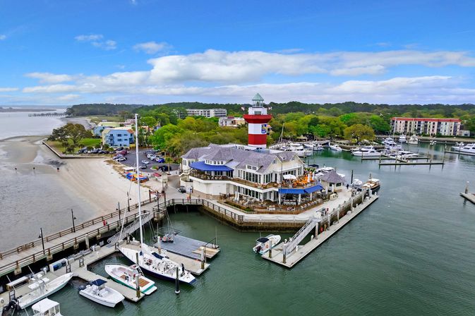 Aerial view of a coastal marina with a red-and-white lighthouse, waterfront restaurant on a pier, docked boats and yachts, sandy beach and tree-lined shoreline under a blue sky.