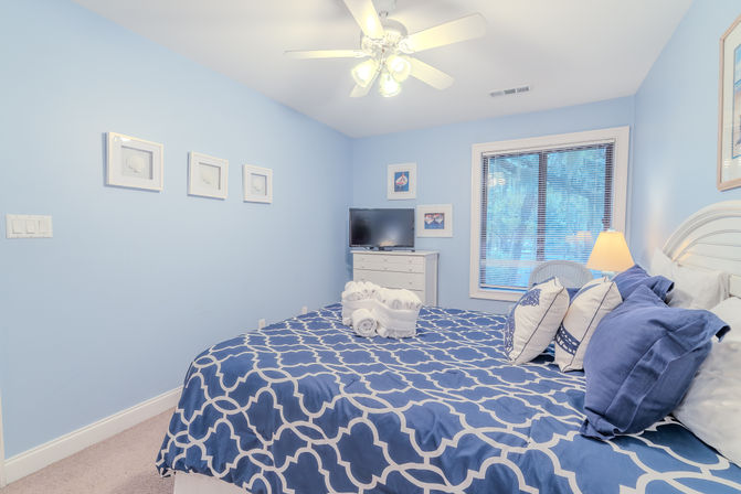 Bright coastal bedroom with pale blue walls, king bed in navy geometric duvet and decorative pillows, folded white towels on the bed, white dresser with TV, window blinds, ceiling fan and bedside lamp