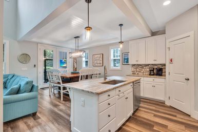 Open-concept coastal-inspired kitchen and dining area with white cabinets, marble-look island and sink, pendant lighting, wood plank floors, teal sofa, and glass patio doors.
