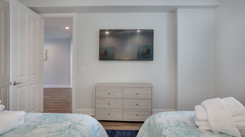 Coastal-style twin bedroom with two beds dressed in seaweed-pattern quilts and folded white towels, light wood six-drawer dresser beneath a wall-mounted flat-screen TV, white walls, hardwood floors, and an open door to a hallway.