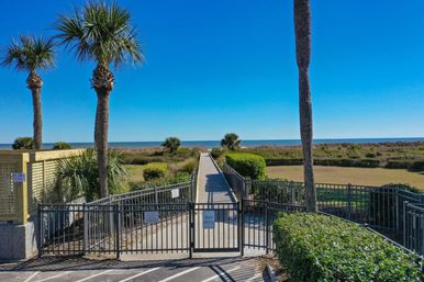 Sunny coastal boardwalk and gated beach access framed by palm trees, leading across dunes to the blue ocean under a clear sky.