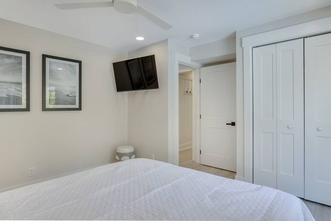 Bright modern guest bedroom with white quilted bed, wall-mounted TV, two framed seascape prints, white sliding closet doors, ceiling fan, and neutral decor