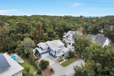 Aerial view of a two-story blue coastal home with white trim, attached garage and front porch on a tree-lined lot near the shore, neighboring backyard pool visible