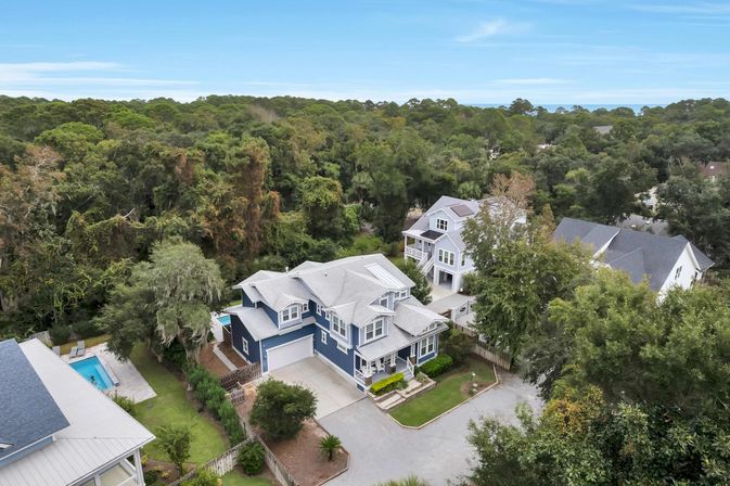 Aerial view of a two-story blue coastal home with white trim, attached garage and front porch on a tree-lined lot near the shore, neighboring backyard pool visible