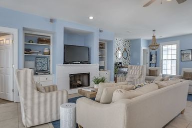 Airy coastal-style living room with pale blue walls, beige sofas and striped armchairs around a wooden coffee table, white fireplace with TV above, built-in shelves and wicker chandelier.
