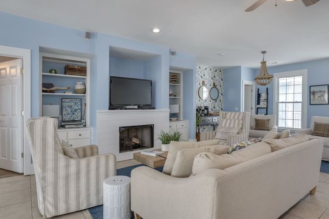 Airy coastal-style living room with pale blue walls, beige sofas and striped armchairs around a wooden coffee table, white fireplace with TV above, built-in shelves and wicker chandelier.