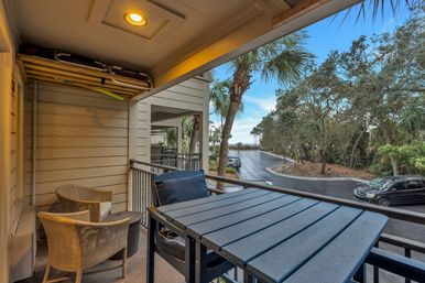 Covered coastal balcony with wicker chairs and navy-cushioned table overlooking palm-lined driveway and distant ocean view
