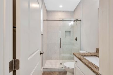 Bright modern residential bathroom with sliding glass walk-in shower and marble tile, white toilet and vanity with granite countertop, recessed ceiling light