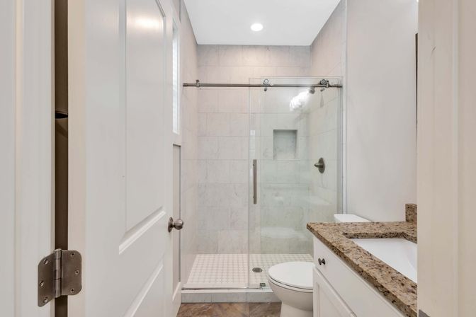 Bright modern residential bathroom with sliding glass walk-in shower and marble tile, white toilet and vanity with granite countertop, recessed ceiling light