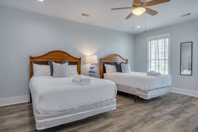 Bright coastal-style guest bedroom with two double beds in white linens, wooden headboards, blue accent pillows, a striped bedside lamp between beds, ceiling fan, shuttered window, and wood-look flooring