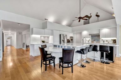 Bright modern open-concept kitchen with vaulted ceiling and fan, white cabinetry, large glossy island with black bar stools, stainless steel appliances, light hardwood floors and decorative metal heron sculptures