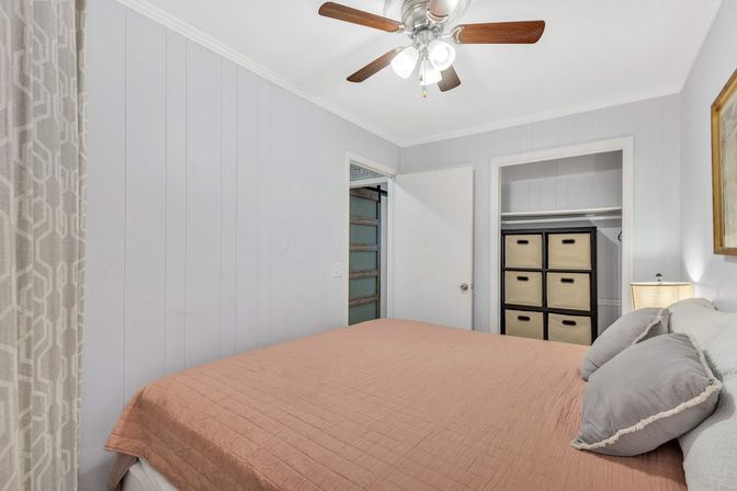 Cozy light-gray bedroom with a ceiling fan, blush quilted bed, patterned curtains, bedside lamp and open closet showing stacked cube storage bins — neutral modern decor.