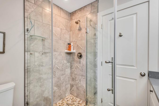 Sleek glass-enclosed shower in a modern bathroom with gray marble-look wall tiles, pebble-stone floor, chrome showerhead and handle, corner shelf with shampoo bottles, and a white paneled door