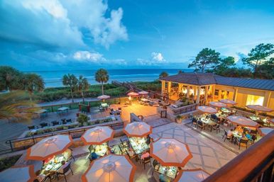 Oceanfront restaurant patio at twilight with lit umbrella tables, wooden boardwalk to sandy beach, palm trees and calm blue sea — seaside dining with sunset views.